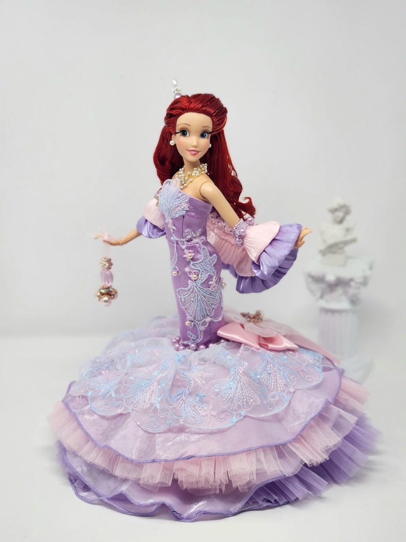 Op de afbeelding: Een prinsessenpop met rood haar, gekleed in een lavendelkleurige jurk met gedetailleerde borduursels en een gelaagde rok. De jurk heeft roze en blauwe accenten en de pop is versierd met sieraden.