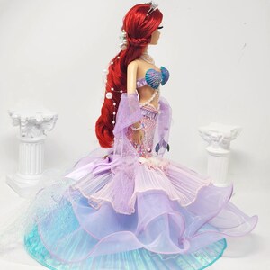 May include: A doll with long red hair, wearing a mermaid-inspired gown. The gown features a seashell bra, a layered skirt in shades of purple, blue, and pink, and a pearl necklace. The doll has a tiara and is posed against a white background.