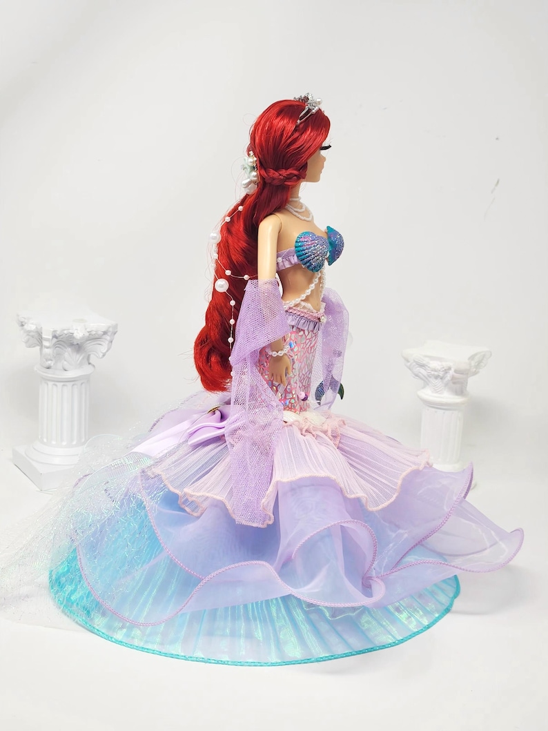 May include: A doll with long red hair, wearing a mermaid-inspired gown. The gown features a seashell bra, a layered skirt in shades of purple, blue, and pink, and a pearl necklace. The doll has a tiara and is posed against a white background.