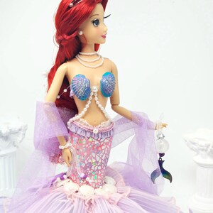 May include: A doll of a mermaid with red hair, wearing a seashell bra and a shimmering pink and purple mermaid tail. The doll has a pearl necklace and a silver tiara. The doll is holding a small, decorative item.