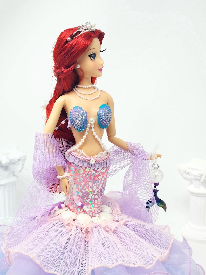 May include: A doll of a mermaid with red hair, wearing a seashell bra and a shimmering pink and purple mermaid tail. The doll has a pearl necklace and a silver tiara. The doll is holding a small, decorative item.