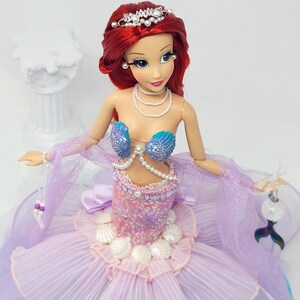 May include: A doll of a mermaid princess with red hair, wearing a seashell bra and a lavender and pink dress. The doll has a pearl necklace, a tiara, and a decorative tail. The doll is holding a decorative item.