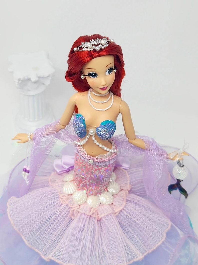 May include: A doll of a mermaid princess with red hair, wearing a seashell bra and a lavender and pink dress. The doll has a pearl necklace, a tiara, and a decorative tail. The doll is holding a decorative item.