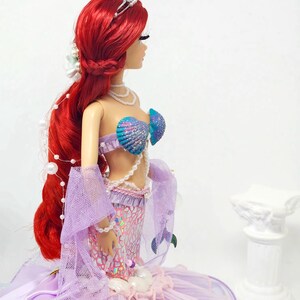 May include: A doll dressed as a mermaid, featuring vibrant red hair, a seashell bra, and a shimmering purple and pink tail. The doll wears a silver tiara and pearl necklaces, with a flowing lavender skirt.