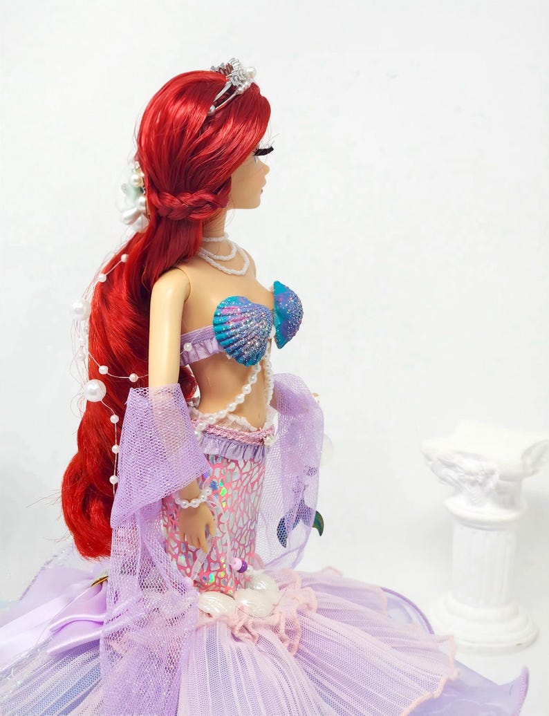 May include: A doll dressed as a mermaid, featuring vibrant red hair, a seashell bra, and a shimmering purple and pink tail. The doll wears a silver tiara and pearl necklaces, with a flowing lavender skirt.