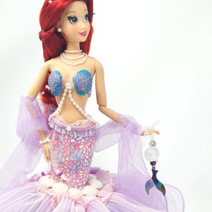 May include: A doll of a mermaid with red hair, wearing a seashell bra and a pink and purple scale-patterned skirt. The doll has a tiara and pearl necklace. She holds a clear orb with a mermaid tail charm.
