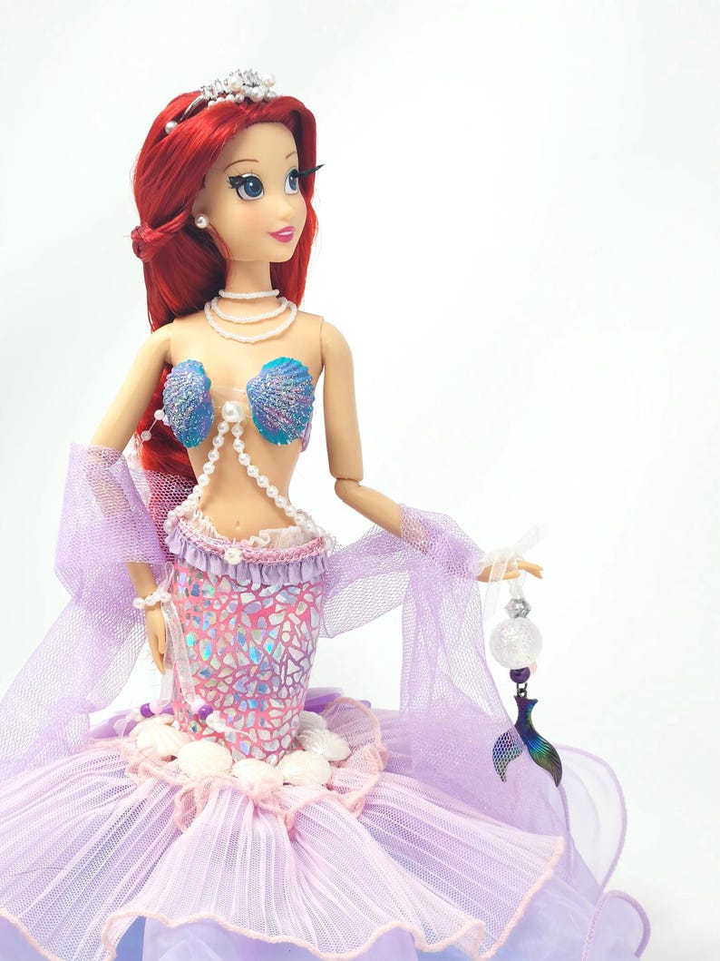 May include: A doll of a mermaid with red hair, wearing a seashell bra and a pink and purple scale-patterned skirt. The doll has a tiara and pearl necklace. She holds a clear orb with a mermaid tail charm.