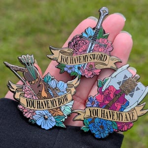 May include: Three enamel pins with floral designs and banners. One pin features a sword, another a bow and arrow, and the third an axe. Each banner reads "YOU HAVE MY..." followed by the item.