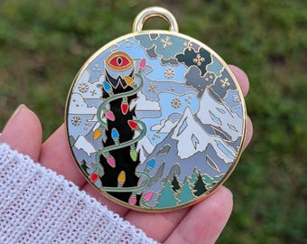 May include: A circular gold-rimmed enamel pin featuring a black tower with a single red eye at the top, wrapped in colorful holiday lights. The background depicts snowy mountains, a light blue sky, and falling snowflakes in a winter landscape.