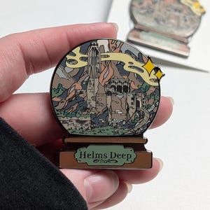 Puede incluir: Un pin de esmalte dorado en forma de bola de nieve con una representaci&oacute;n de Helm's Deep del Se&ntilde;or de los Anillos. El pin presenta una ilustraci&oacute;n detallada de la fortaleza, las monta&ntilde;as y una nube de humo arremolinada. La base del pin dice "Helms Deep".