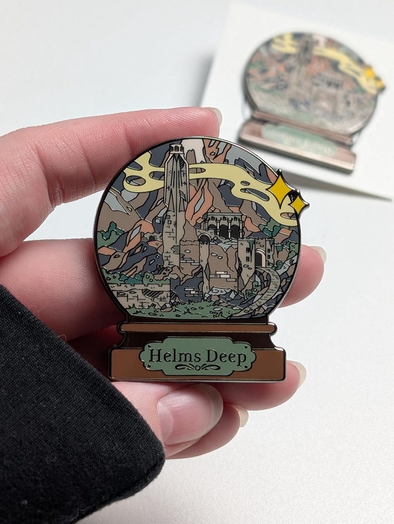 Puede incluir: Un pin de esmalte dorado en forma de bola de nieve con una representaci&oacute;n de Helm's Deep del Se&ntilde;or de los Anillos. El pin presenta una ilustraci&oacute;n detallada de la fortaleza, las monta&ntilde;as y una nube de humo arremolinada. La base del pin dice "Helms Deep".