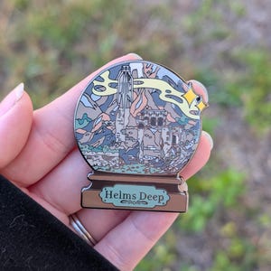 Puede incluir: Un pin de esmalte plateado con forma de bola de nieve con una representaci&oacute;n de Helm's Deep del Se&ntilde;or de los Anillos. El pin tiene una base marr&oacute;n con las palabras "Helms Deep" escritas en blanco.
