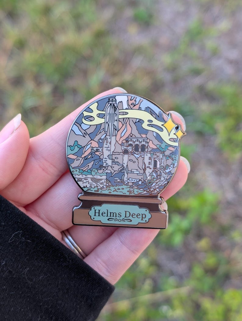 Puede incluir: Un pin de esmalte plateado con forma de bola de nieve con una representaci&oacute;n de Helm's Deep del Se&ntilde;or de los Anillos. El pin tiene una base marr&oacute;n con las palabras "Helms Deep" escritas en blanco.