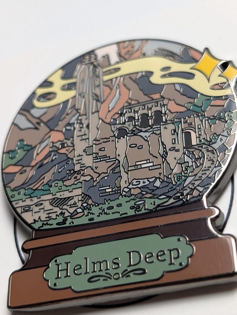 Puede incluir: Un pin de esmalte plateado con forma de bola de nieve con una representaci&oacute;n de Helm's Deep, una fortaleza de la saga de El Se&ntilde;or de los Anillos, en su interior. La base de la bola de nieve es marr&oacute;n con un letrero verde que dice "Helms Deep".