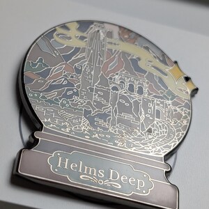 Puede incluir: Un pin de esmalte plateado en forma de bola de nieve con una ilustraci&oacute;n detallada de Helm's Deep del Se&ntilde;or de los Anillos. El pin tiene un esquema de color azul claro, gris y amarillo.