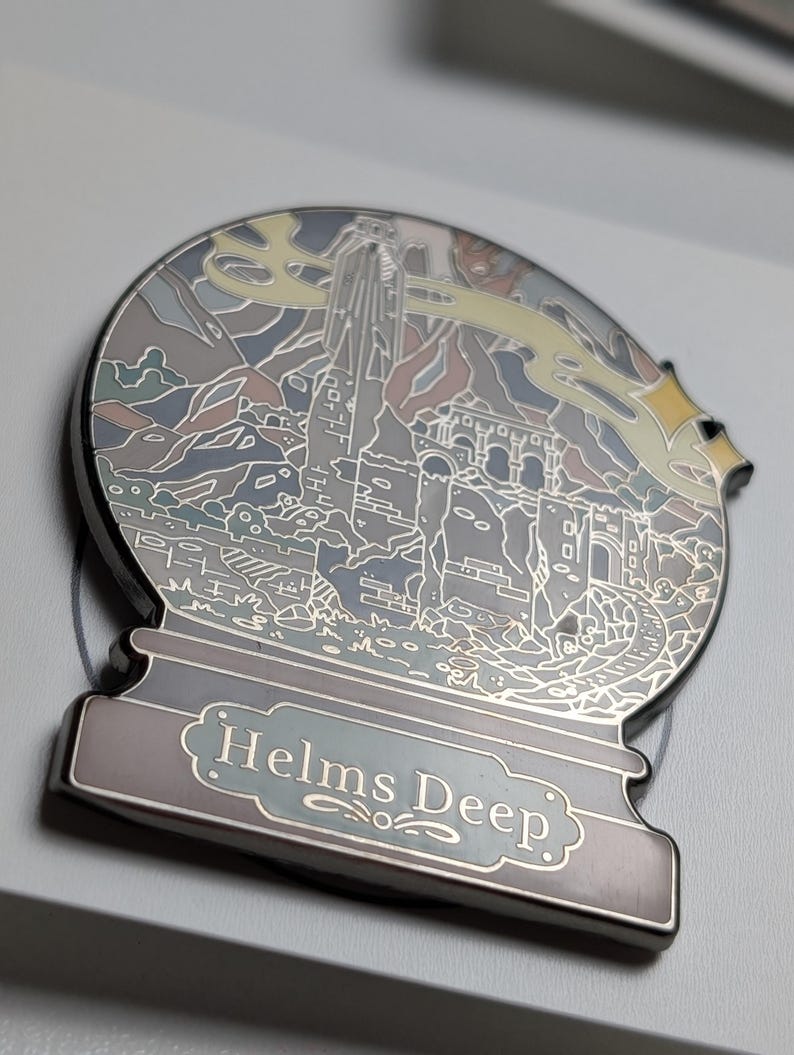 Puede incluir: Un pin de esmalte plateado en forma de bola de nieve con una ilustraci&oacute;n detallada de Helm's Deep del Se&ntilde;or de los Anillos. El pin tiene un esquema de color azul claro, gris y amarillo.