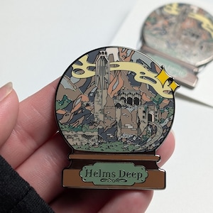 Puede incluir: Un pin de esmalte plateado con forma de bola de nieve con una ilustraci&oacute;n detallada de Helm's Deep de El Se&ntilde;or de los Anillos. El pin tiene una base marr&oacute;n con las palabras "Helms Deep" escritas en verde.