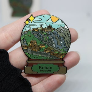 Peut inclure: Une &eacute;pingle en &eacute;mail dor&eacute;e en forme de boule &agrave; neige repr&eacute;sentant un village de montagne avec un ch&acirc;teau au sommet. La boule &agrave; neige a une base verte avec le mot "Rohan" inscrit dessus.