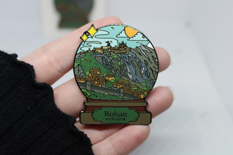 Peut inclure: Une &eacute;pingle en &eacute;mail dor&eacute;e en forme de boule &agrave; neige repr&eacute;sentant un village de montagne avec un ch&acirc;teau au sommet. La boule &agrave; neige a une base verte avec le mot "Rohan" inscrit dessus.