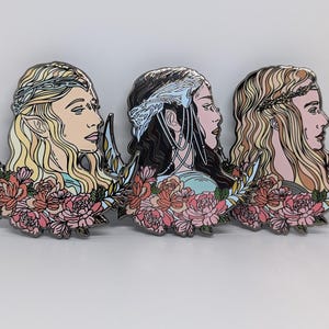 May include: Three enamel pins featuring profile portraits of women with long hair, floral accents, and crescent moon shapes. The pins have a color palette of pink, cream, black, and gold. The pins are likely fantasy-themed.