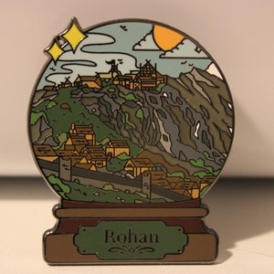 Peut inclure: Une &eacute;pingle en m&eacute;tal argent&eacute; en forme de boule &agrave; neige repr&eacute;sentant un village de montagne avec un sch&eacute;ma de couleurs vert, marron et gris. La boule &agrave; neige a une base marron avec une pancarte verte sur laquelle est inscrit "Rohan".