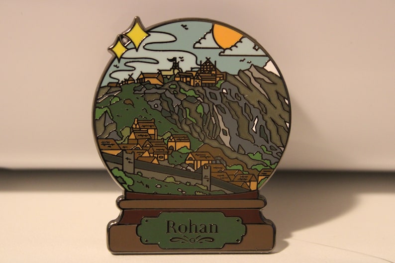 Peut inclure: Une &eacute;pingle en m&eacute;tal argent&eacute; en forme de boule &agrave; neige repr&eacute;sentant un village de montagne avec un sch&eacute;ma de couleurs vert, marron et gris. La boule &agrave; neige a une base marron avec une pancarte verte sur laquelle est inscrit "Rohan".