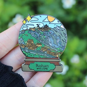 Peut inclure: Une &eacute;pingle en &eacute;mail en forme de globe &agrave; neige argent&eacute; avec une illustration verte, bleue et marron d'un village de montagne avec le texte "Rohan" sur la base.
