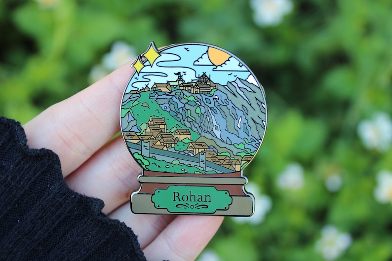 Peut inclure: Une &eacute;pingle en &eacute;mail en forme de globe &agrave; neige argent&eacute; avec une illustration verte, bleue et marron d'un village de montagne avec le texte "Rohan" sur la base.
