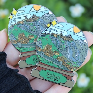 Peut inclure: Deux &eacute;pingles en &eacute;mail argent&eacute; en forme de boules &agrave; neige, repr&eacute;sentant un paysage montagneux avec un village et un ch&acirc;teau. La base de chaque &eacute;pingle est brune avec une pancarte verte qui indique "Rohan".