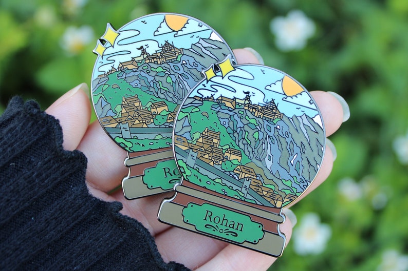 Peut inclure: Deux &eacute;pingles en &eacute;mail argent&eacute; en forme de boules &agrave; neige, repr&eacute;sentant un paysage montagneux avec un village et un ch&acirc;teau. La base de chaque &eacute;pingle est brune avec une pancarte verte qui indique "Rohan".