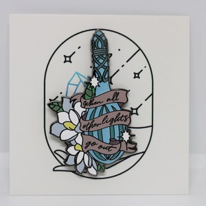 May include: A blue enamel pin with a floral design and a banner that reads "When all other lights go out".