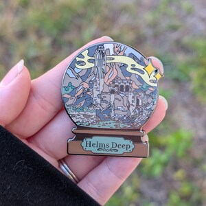 Puede incluir: Un pin de esmalte plateado con forma de bola de nieve con una representaci&oacute;n de Helm's Deep del Se&ntilde;or de los Anillos. El pin presenta una ilustraci&oacute;n detallada de la fortaleza, las monta&ntilde;as y una estrella amarilla.