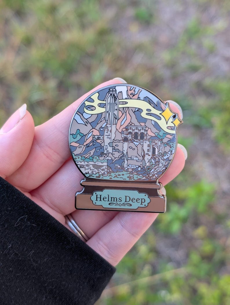 Puede incluir: Un pin de esmalte plateado con forma de bola de nieve con una representaci&oacute;n de Helm's Deep del Se&ntilde;or de los Anillos. El pin presenta una ilustraci&oacute;n detallada de la fortaleza, las monta&ntilde;as y una estrella amarilla.