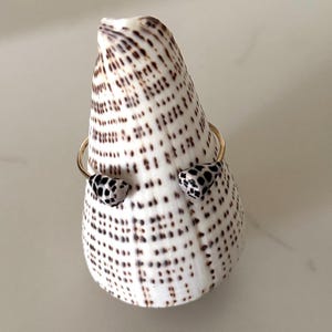 May include: A gold-toned bracelet with black and white spotted accents. The bracelet is displayed on a large, white seashell with brown spots. The seashell is upright, and the bracelet is positioned in front of it.