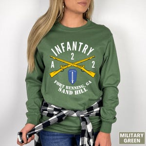 May include: Military green long-sleeve shirt with a graphic that reads "INFANTRY 2 A 2" above crossed yellow rifles. Below the rifles is a shield with a blue and white design, and the text "FORT BENNING, GA SAND HILL".