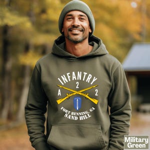 May include: Military green hoodie with a white and yellow graphic. The graphic includes the word "INFANTRY", crossed rifles, and the text "FORT BENNING, GA SAND HILL". The hoodie has a front pocket and a drawstring hood. The text "Military Green" is in the bottom right corner.