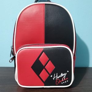 May include: A mini backpack featuring a split red and black color scheme with white piping. The front pocket displays two red diamond shapes on a black background and the text Harley Quinn xoxo in white and red script with small heart accents.