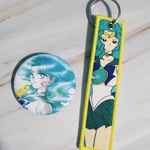 May include: A round button and a rectangular keychain featuring a teal-haired anime character. The button has a white background with the character's face. The keychain has a yellow border and a full-body image of the character in a sailor uniform.