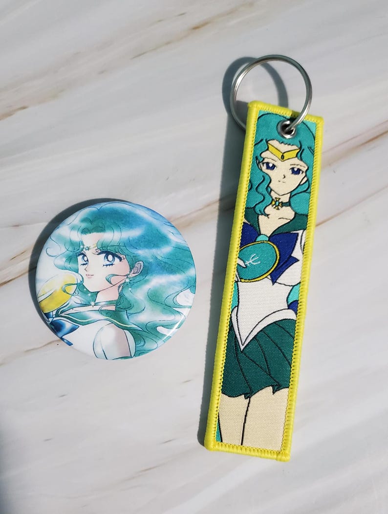 May include: A round button and a rectangular keychain featuring a teal-haired anime character. The button has a white background with the character's face. The keychain has a yellow border and a full-body image of the character in a sailor uniform.