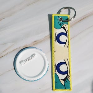 May include: A button and a keychain. The button is round with a white center and a blue and white patterned edge. The keychain is rectangular with a yellow border and features a cartoon eye design with blue and black details.