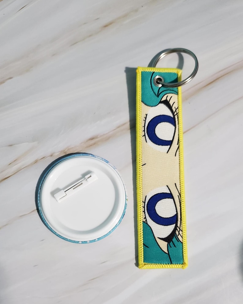 May include: A button and a keychain. The button is round with a white center and a blue and white patterned edge. The keychain is rectangular with a yellow border and features a cartoon eye design with blue and black details.