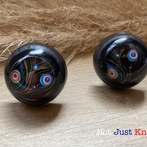 May include: Two black round cabinet knobs with colourful abstract designs. The designs feature swirling lines and circles in shades of red, blue, green, and yellow on a black background. The knobs are shown on a wooden surface.
