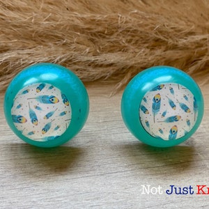 May include: Two turquoise round cabinet knobs with a white background and a blue and yellow feather pattern. The knobs are on a wooden surface.