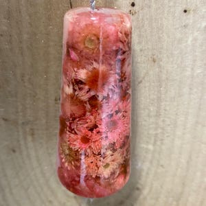 May include: A clear, cylindrical object filled with dried pink and peach-coloured flowers. A silver-coloured string is attached to the top. The object is set against a wooden background.