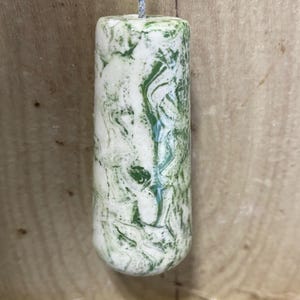 May include: A cylindrical light pull with a green and white marbled design. The pull is suspended by a silver cord. The background is a light brown wooden surface.