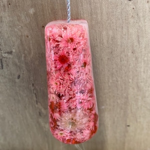 May include: A rectangular resin pendant with rounded bottom edges, filled with numerous small, dried pink flowers. The clear, glossy material encases the floral arrangement, and a thin, silver-coloured twisted cord is attached to the top centre.