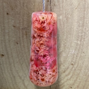 May include: A rectangular pendant made of clear resin containing numerous small, dried pink flowers. The pendant has rounded bottom corners and is suspended from a thin, silver-coloured metallic cord that extends from the top centre.