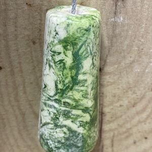 May include: A cylindrical pendant with a tapered shape featuring a marbled pattern of white and various shades of green. The object has a smooth, polished finish and is suspended by a thin, silver-coloured metallic cord threaded through the centre.