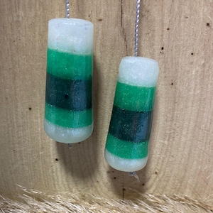 May include: Two cylindrical light pulls, handmade with a colour scheme of white, green, and dark green. Each pull is suspended by a silver-coloured string. The pulls are set against a wooden background.