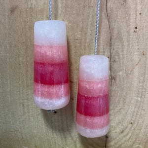 May include: Two handmade, layered candles with a gradient of white, pink, and red hues. Each candle is cylindrical with a tapered shape and a silver string for hanging. The candles are displayed against a wooden background.